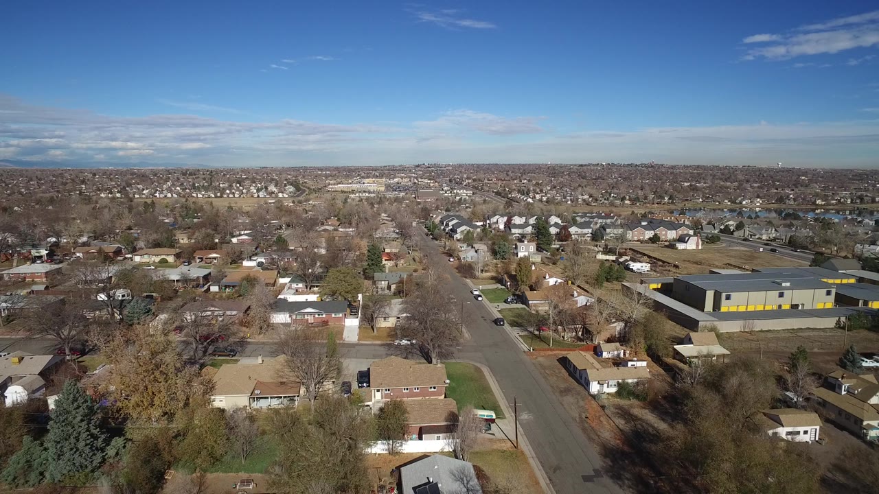 High aerial frame in Westminster, Colorado with rows of houses, a broad road, leafless trees, a large gray industrial structure, and distant suburban development, with no visible people.