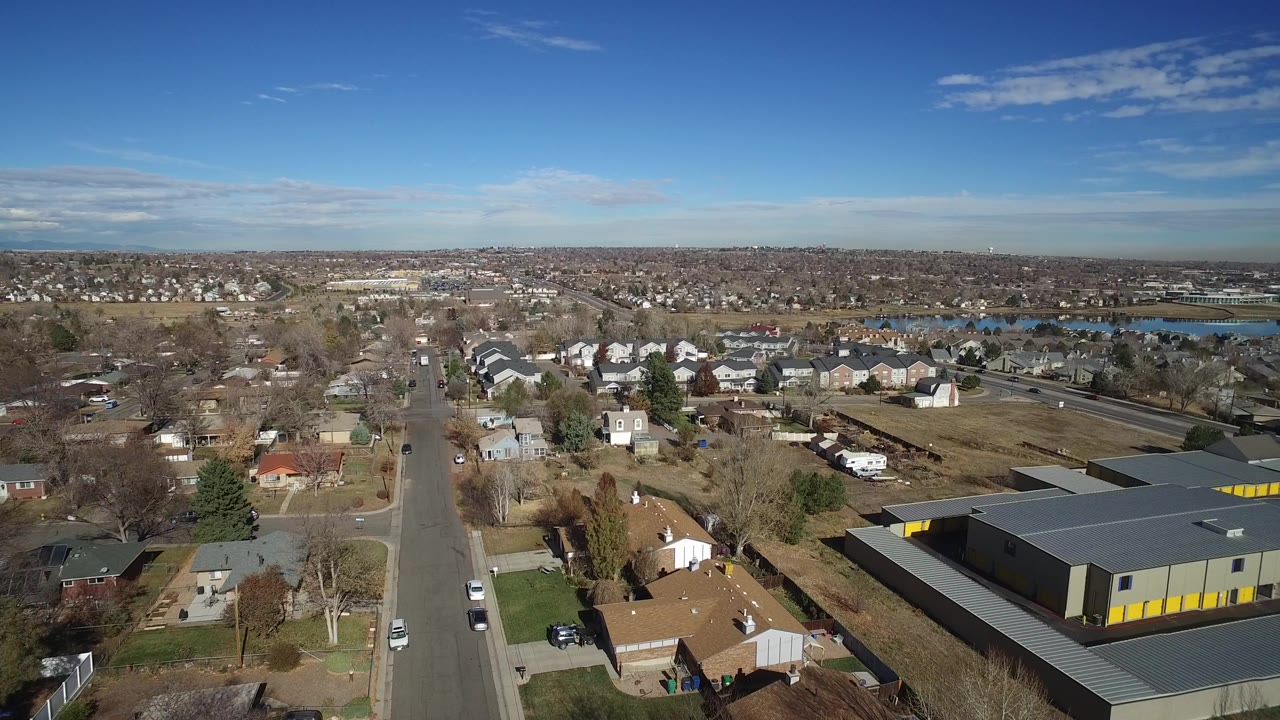 A wide aerial view in Westminster, Colorado shows a residential street, houses, bare trees, a gray building with yellow doors, a distant lake, and no visible people.