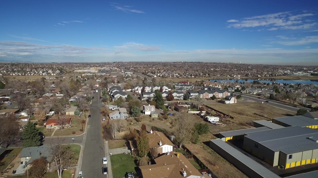 Drone-style view of a neighborhood with a large gray building, houses, roads, and a distant lake.