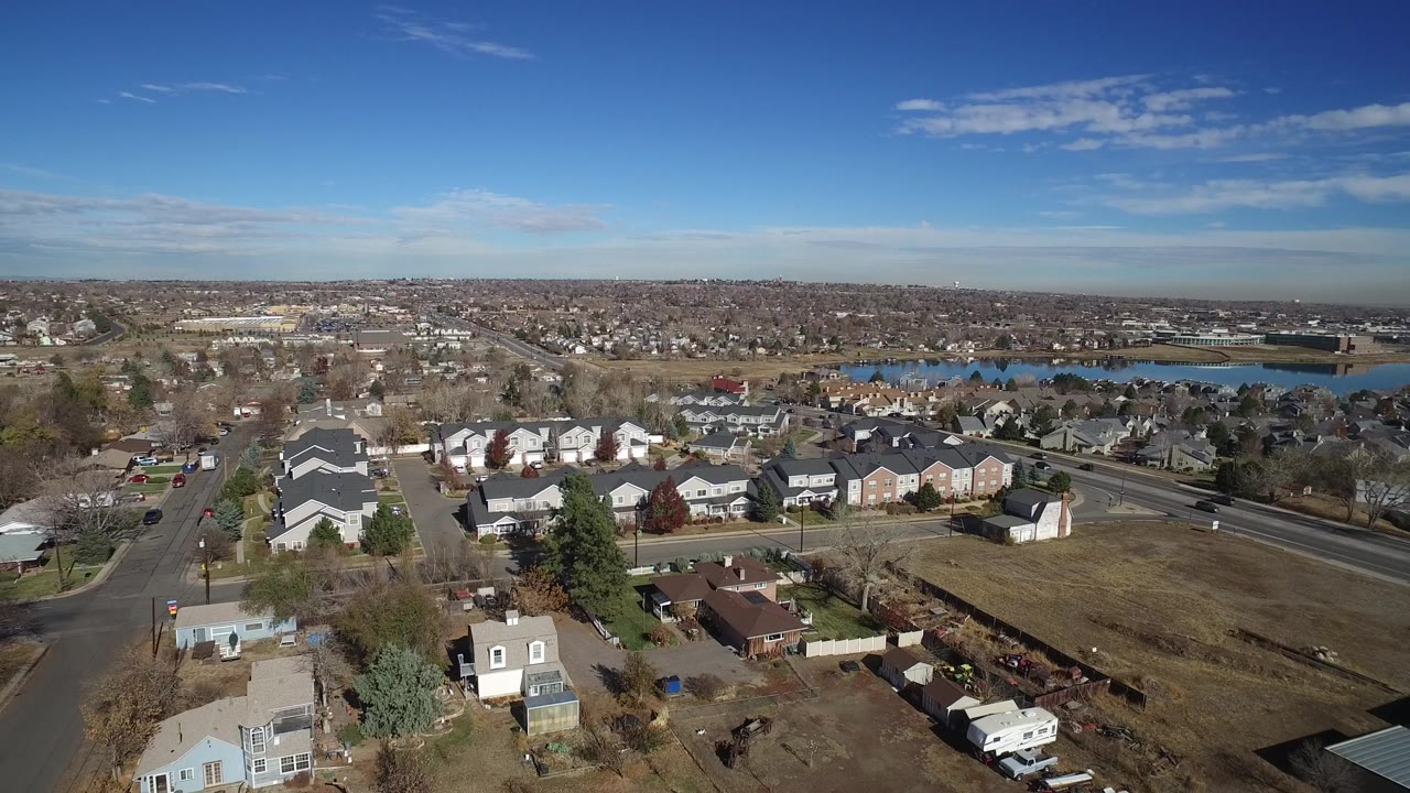 Aerial view over suburban homes, roads, open land, and a distant lake.