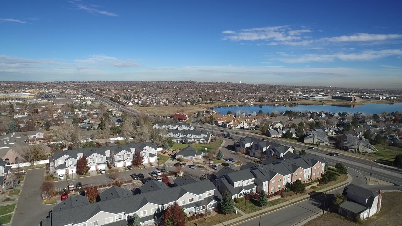 Aerial frame in Westminster, Colorado, looking across gray rooftops and neighborhood streets toward a lake or reservoir, with no visible people.