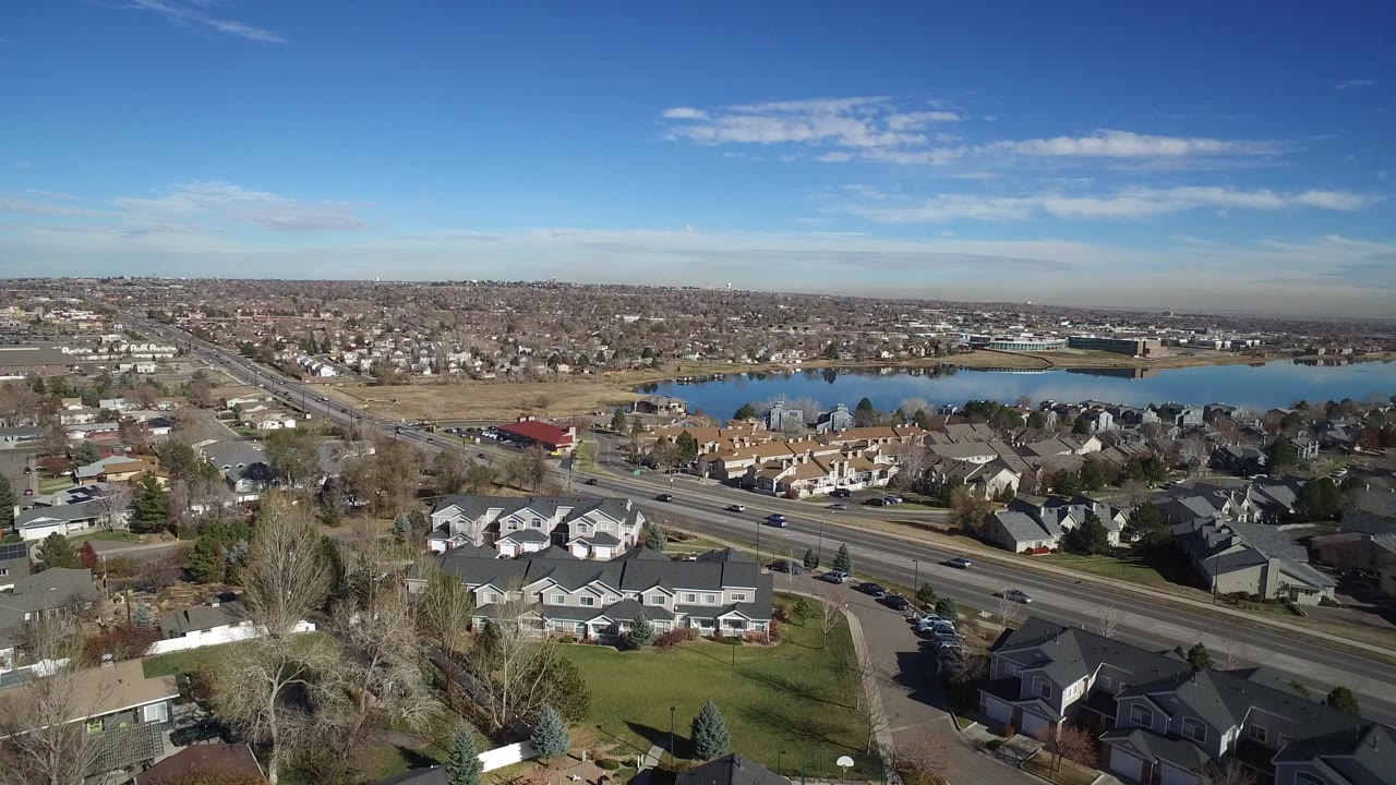 Aerial view over Westminster, Colorado homes with a multi-lane road, small cars, trees, open land, and a lake in the background, with no visible people.
