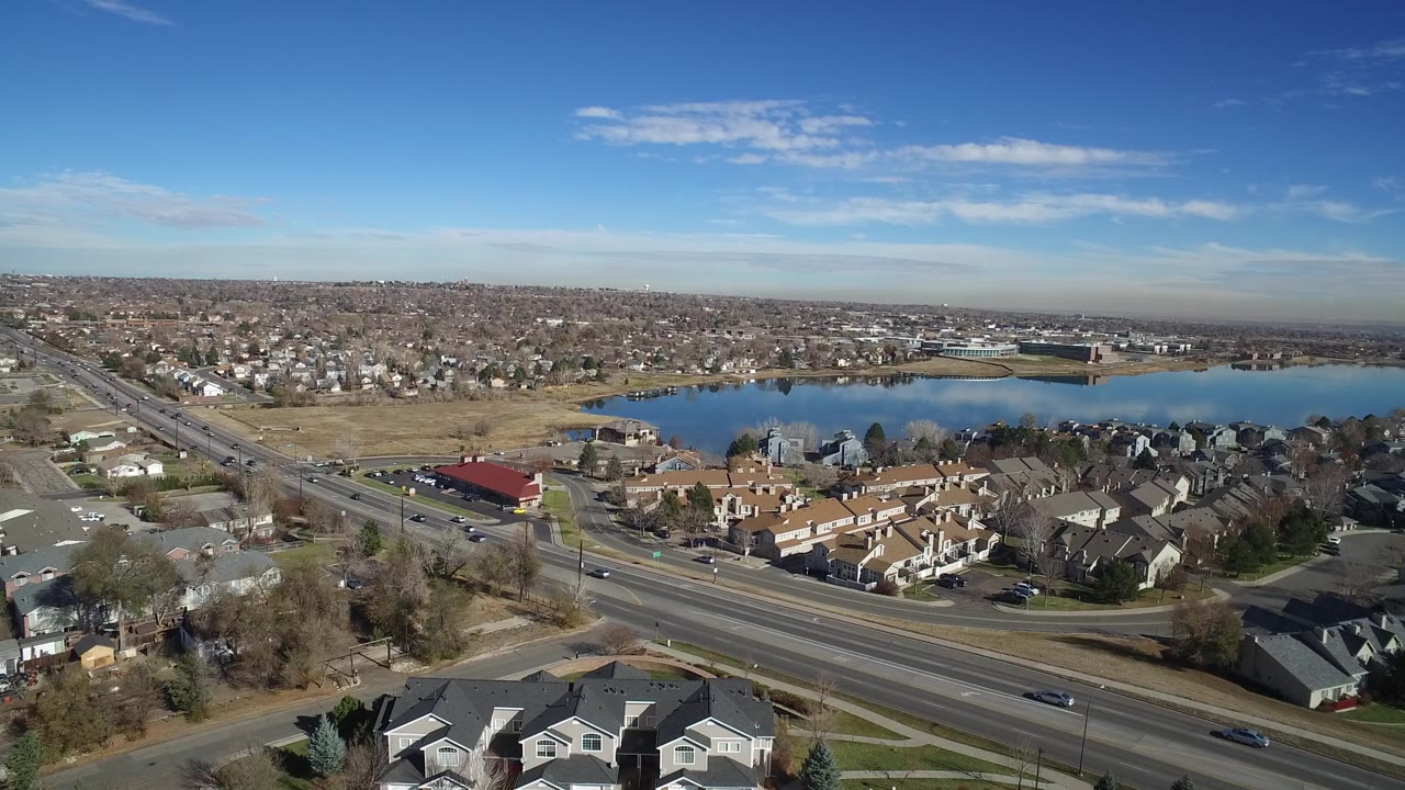 High aerial frame in Westminster, Colorado showing the road corridor angling past residential blocks, a red-roofed building near the lake, distant development, and no visible people.