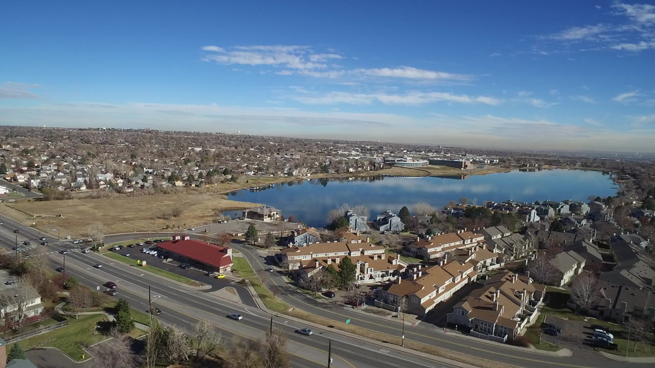 Opening aerial frame over Westminster, Colorado, showing a lake beyond a multi-lane road, tan-roofed homes, dry grass, and no visible people.
