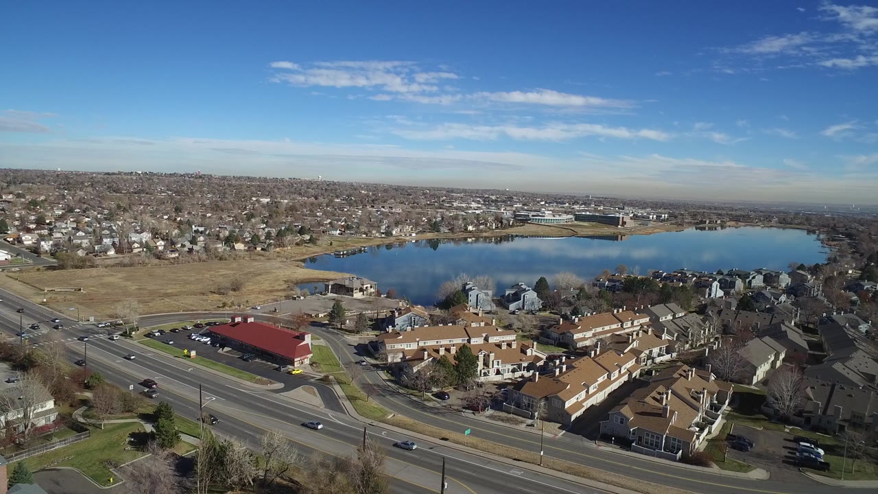Aerial frame in Westminster, Colorado, with the lake centered behind a red-roofed roadside building, rows of homes, moving cars, and no visible people.