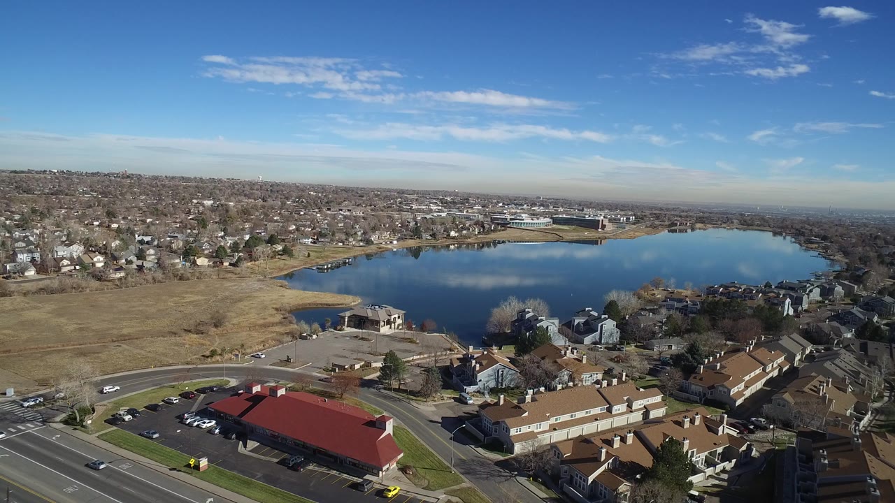 Wide aerial view in Westminster, Colorado showing a small lake behind suburban homes, a red-roofed commercial building near a road, distant neighborhoods, and no visible people.