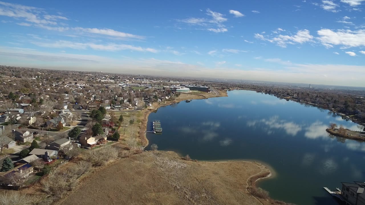 The Westminster, Colorado lake fills the right side of the frame while houses, roads, small docks, leafless trees, and tan open ground remain visible with no visible people.
