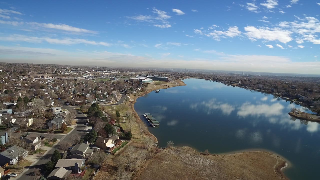 A wide aerial frame over Westminster, Colorado shows private homes and streets along a large blue lake, tan grass in the foreground, cloud reflections on the water, and no visible people.