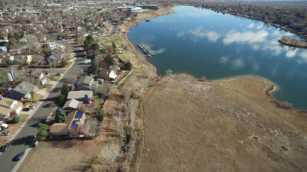 High aerial view in Westminster, Colorado showing suburban houses, a curving street, dry open grass, and a blue lake with cloud reflections, with no visible people.