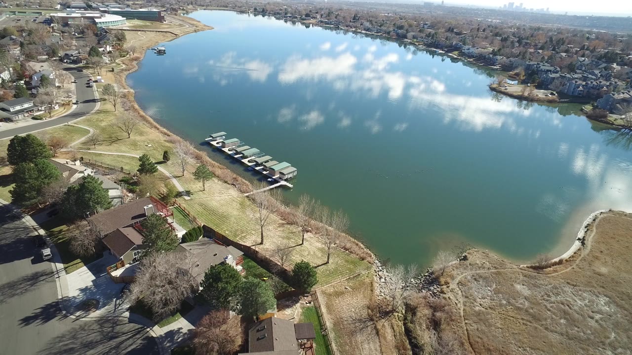 High aerial view in Westminster, Colorado, looking over a blue-green lake with covered moorings, shoreline grass, nearby homes, roads, and cloud reflections on the water, with no clearly visible people.