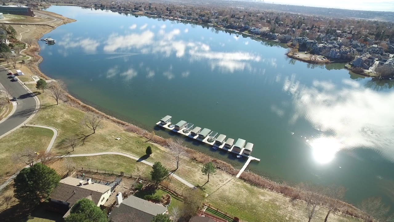 High aerial view over a Westminster, Colorado lake with covered boats at a dock, curved park paths, homes across the water, cloud reflections, and no visible people.