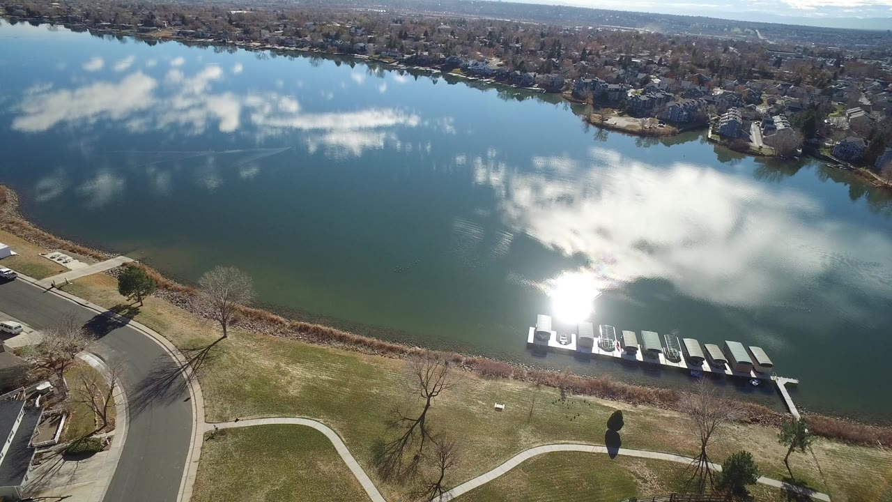 Aerial view over a Westminster, Colorado pond with cloud reflections, bright sun on the water, grassy paths, docks near the near shore, and no visible people.