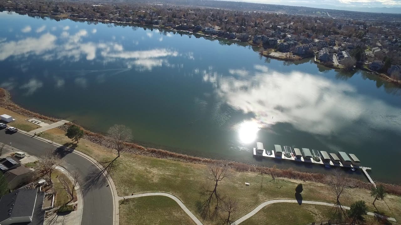 Pond shoreline from above with docks, a road curve, bare trees, and reflected clouds.