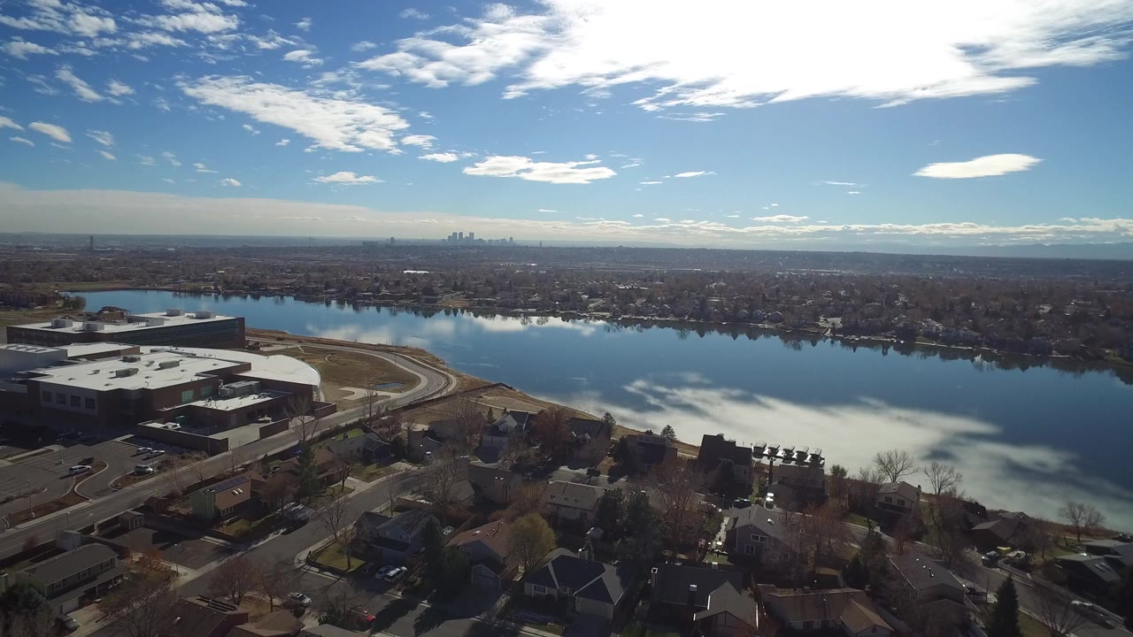 Aerial view over houses beside a reflective lake with a distant city skyline.