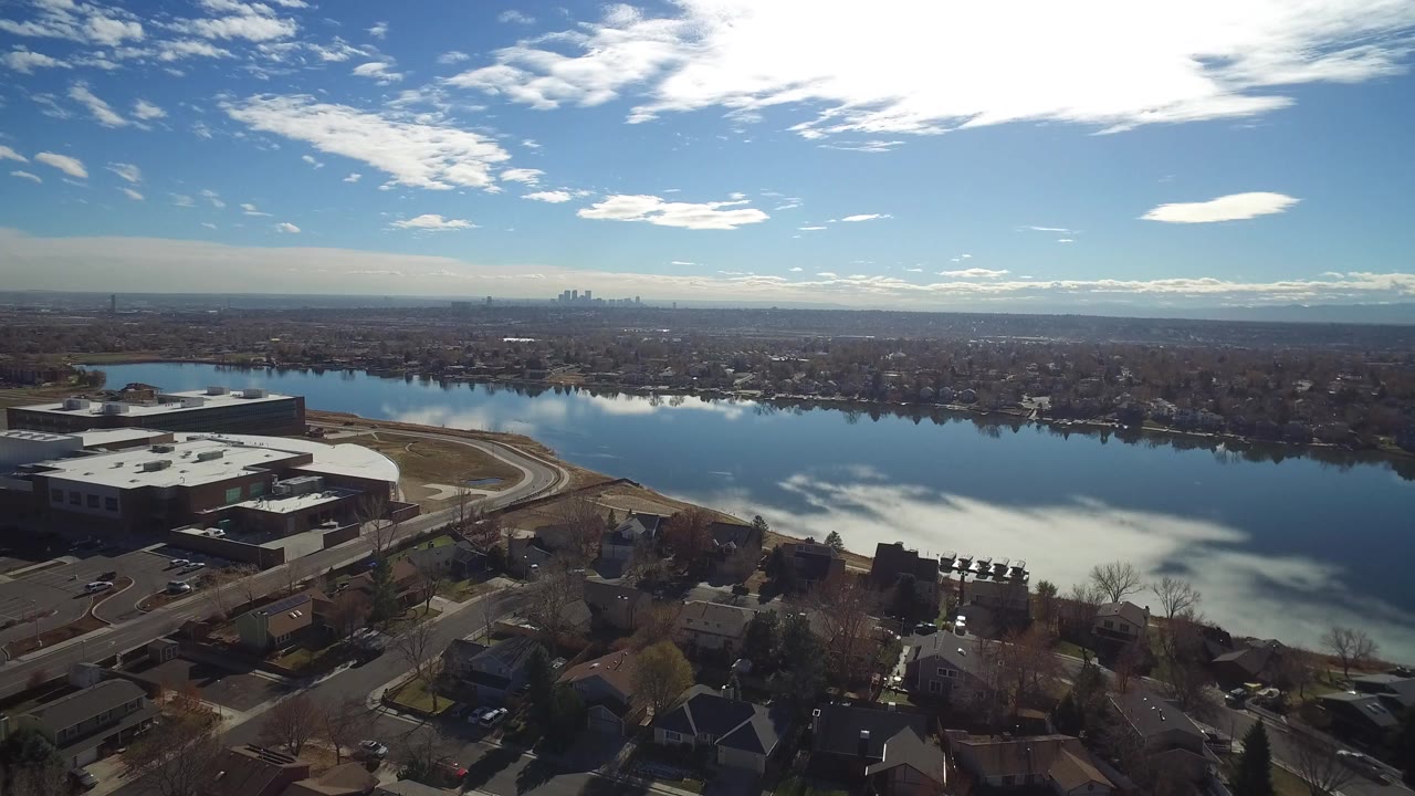 Aerial frame in Westminster, Colorado showing rooftops and a large building in the foreground, lake water across the center, and no visible people.