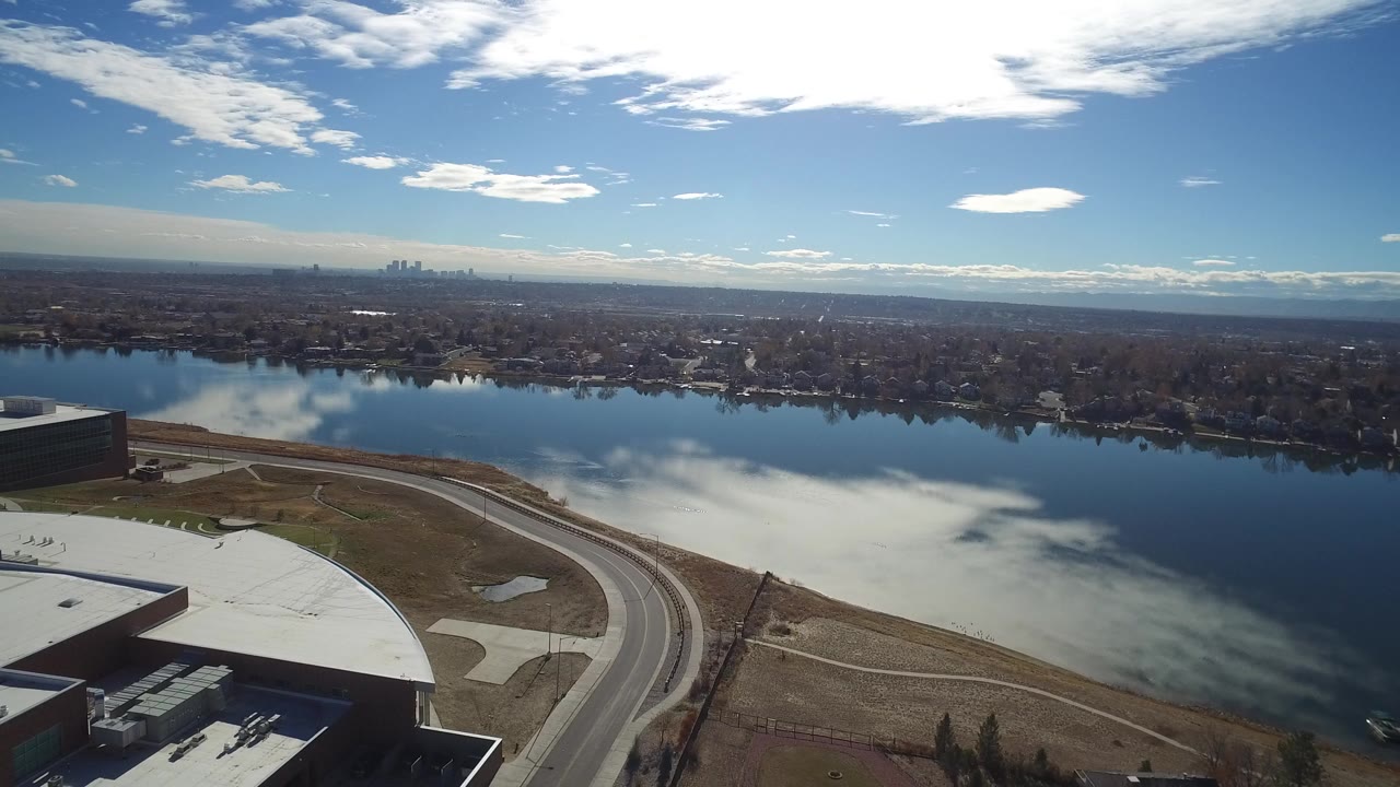 Aerial view of water, curved roads, rooftops, and distant skyline under a bright sky.