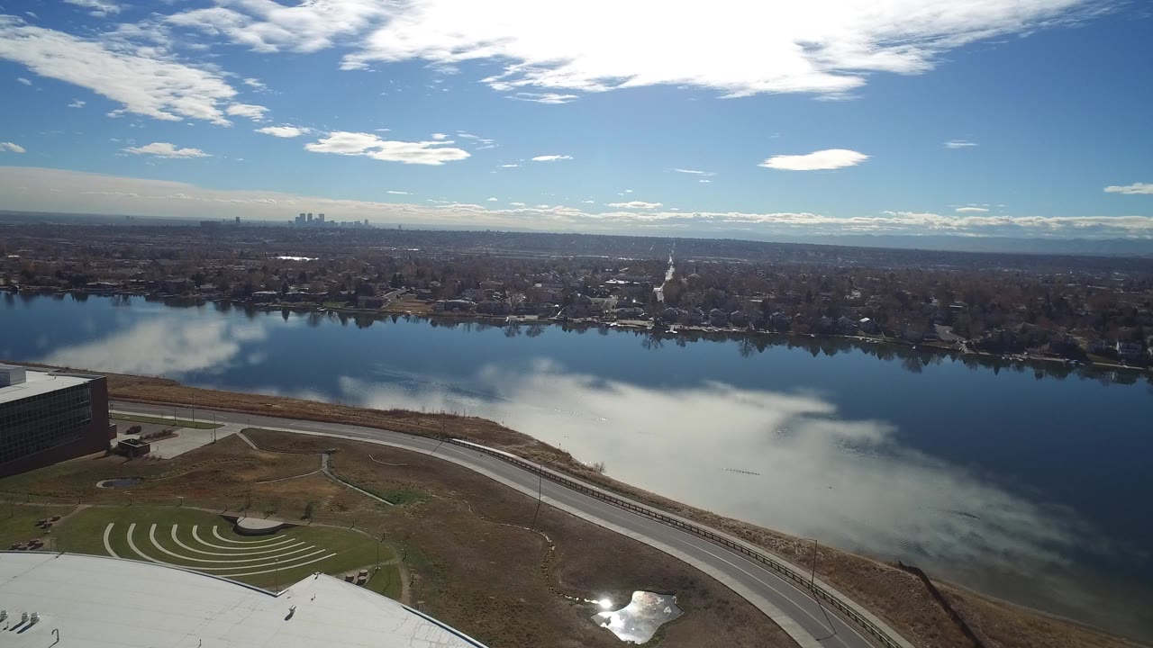 A wide Westminster, Colorado aerial view shows the lake centered under reflected white clouds, foreground road and paths, a partial dark building, distant city skyline, and no visible people.