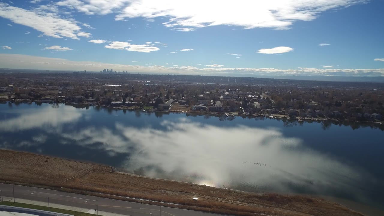 The aerial view continues over the Westminster, Colorado lake, with bright cloud reflections across the water, homes along the far shore, and no visible people.