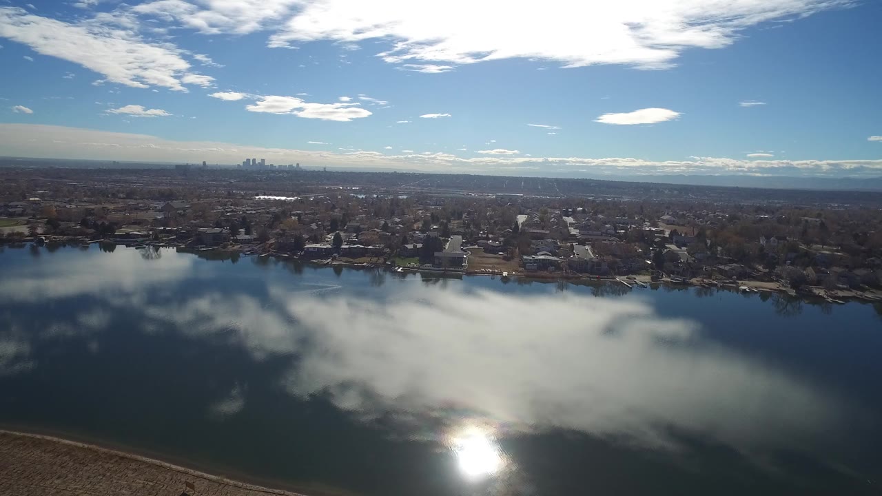 Elevated Westminster, Colorado lake view with a strong sun reflection on the water, houses along the far bank, and the city skyline small on the horizon, with no visible people.