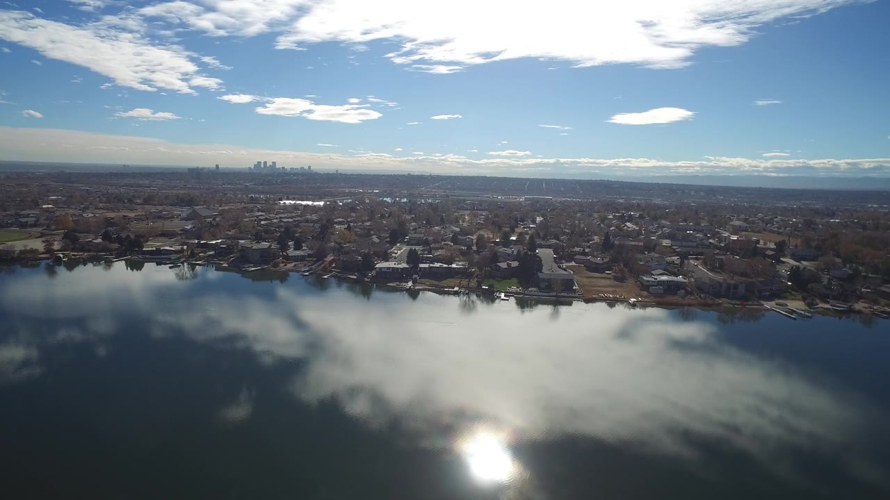 A second Westminster, Colorado aerial frame holds the lake in the foreground with a bright reflection near the lower edge, residential shoreline in the middle distance, distant skyline, and no visible people.