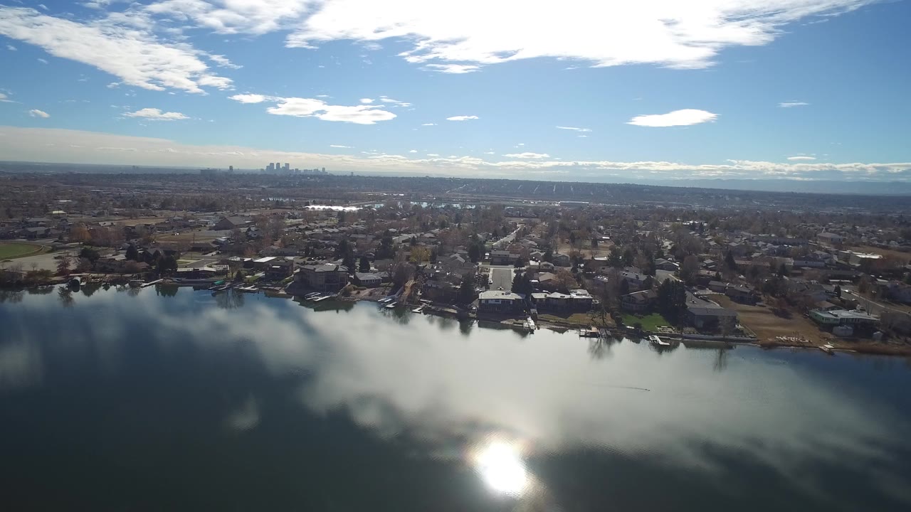 At 0:01, the lake surface reflects bright clouds while Westminster shoreline houses, docks, trees, and a distant city skyline remain visible beyond the water, with no visible people.