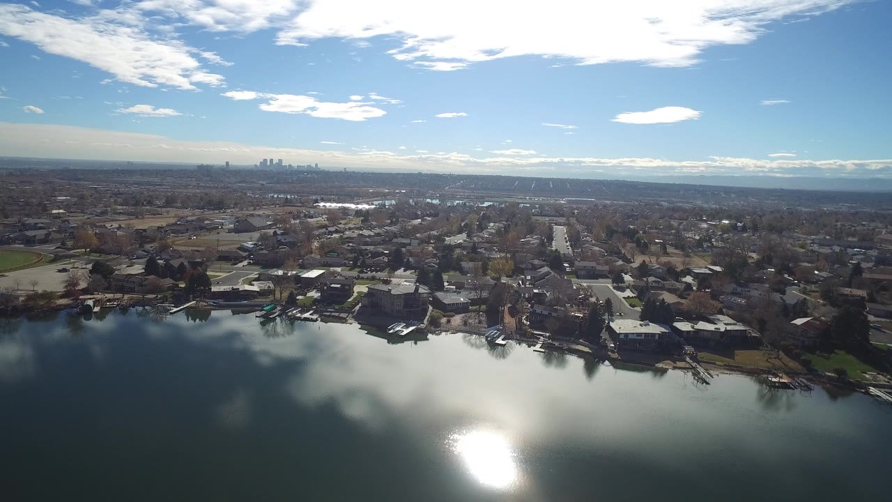 Wide Westminster, Colorado aerial frame with lake glare in the foreground, houses and docks along the shore, gridded streets beyond, distant buildings, and no clearly visible people.