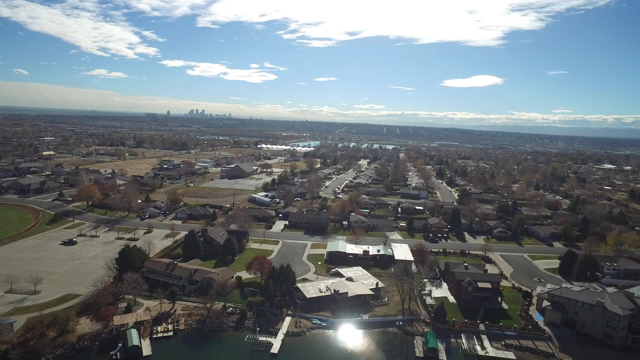 Aerial view of Westminster, Colorado homes, wide streets, bare trees, athletic fields, and distant skyline under scattered clouds, with no visible people.