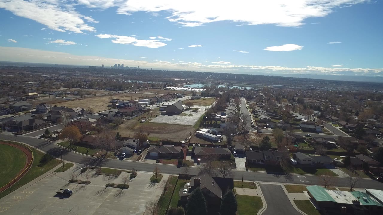 Aerial view over suburban streets, houses, lawns, and distant skyline under blue sky.