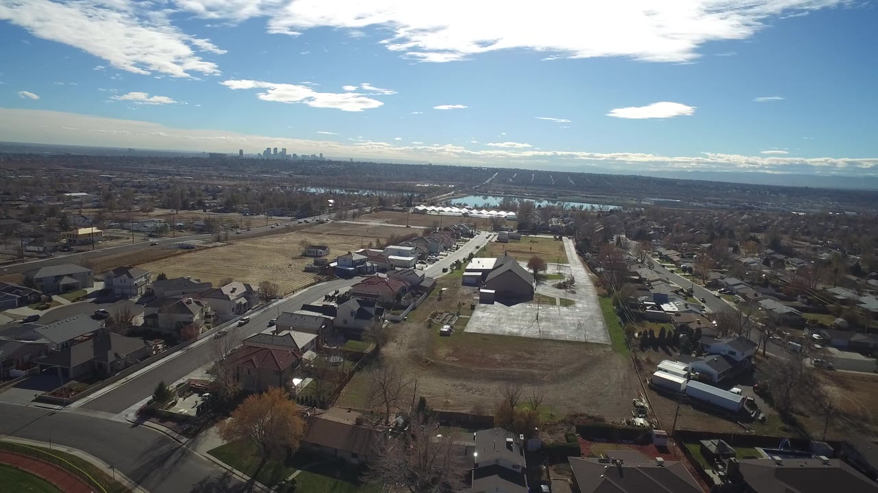 Aerial view in Westminster, Colorado looking across rooftops, paved streets, open brown ground, reflective water, and a distant skyline, with no visible people.
