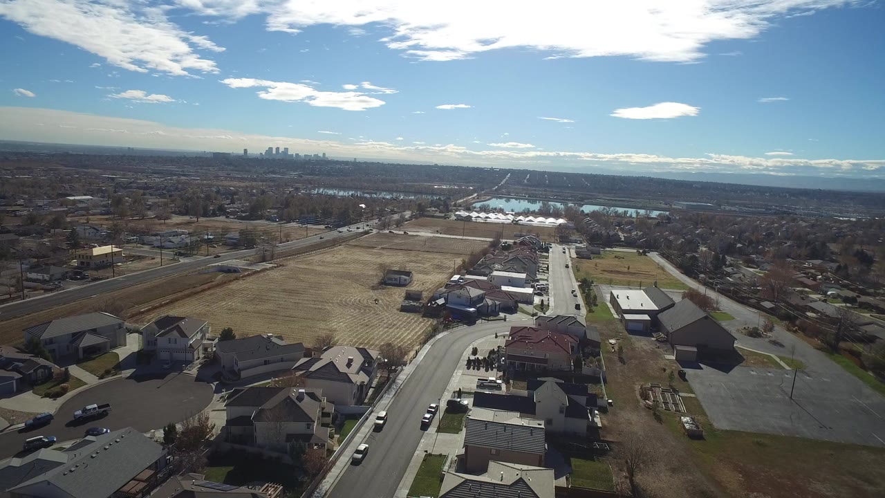 Aerial view over houses, fields, roads, water basins, and a distant skyline.