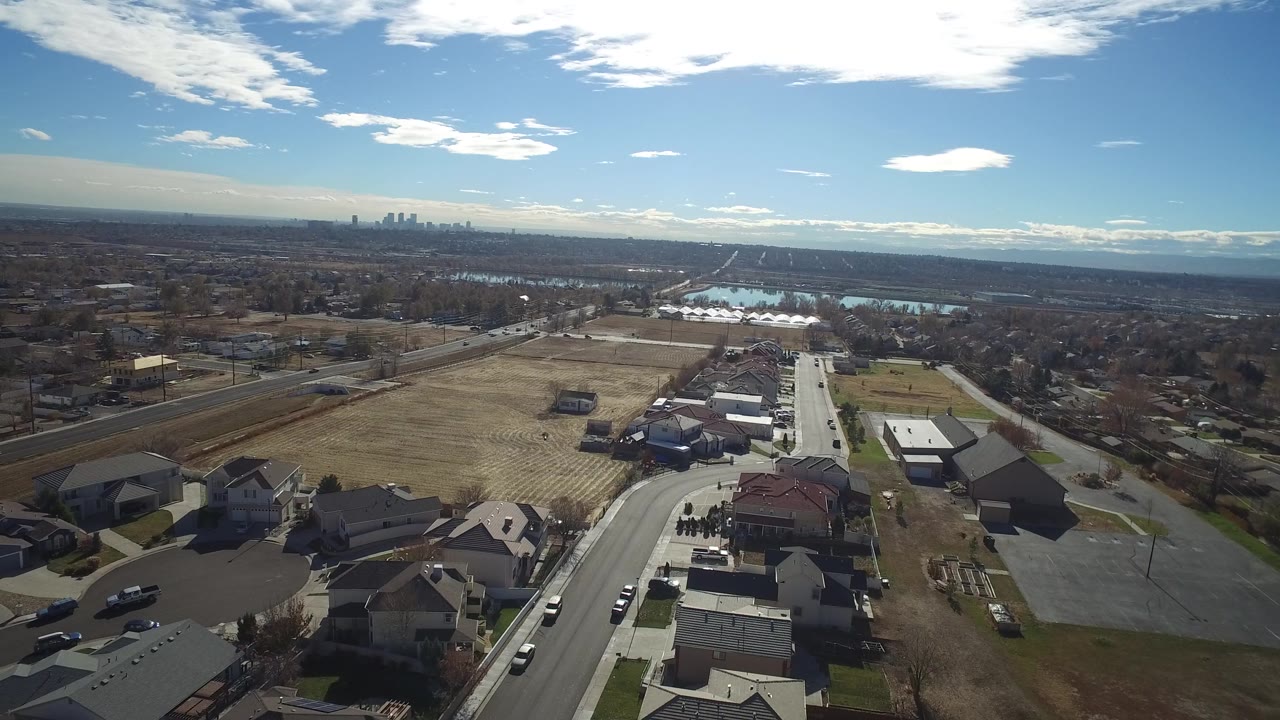 Aerial view in Westminster, Colorado with houses and a cul-de-sac in the foreground, a broad tan field across the center, water basins beyond, distant skyline, and no visible people.