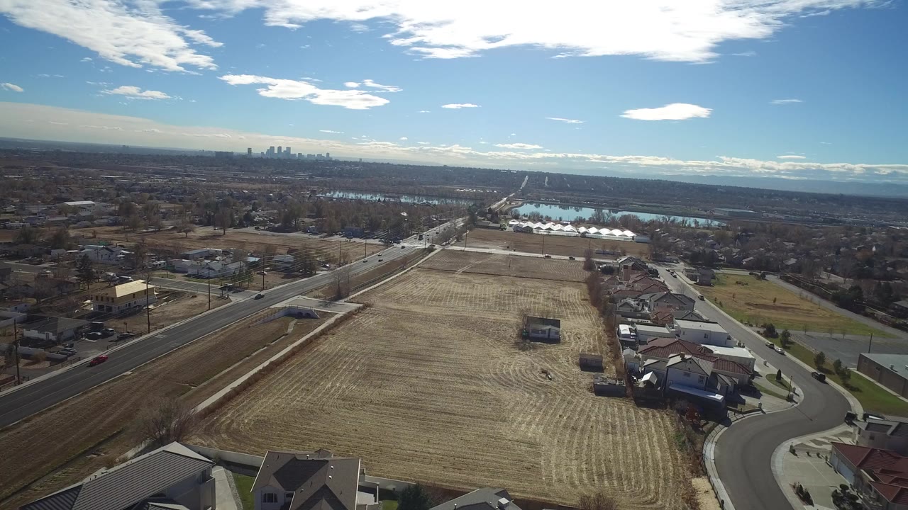 Opening aerial frame over Westminster, Colorado with tan fields in front, houses and a road below, a bright water body beyond, distant skyline on the horizon, and no visible people.