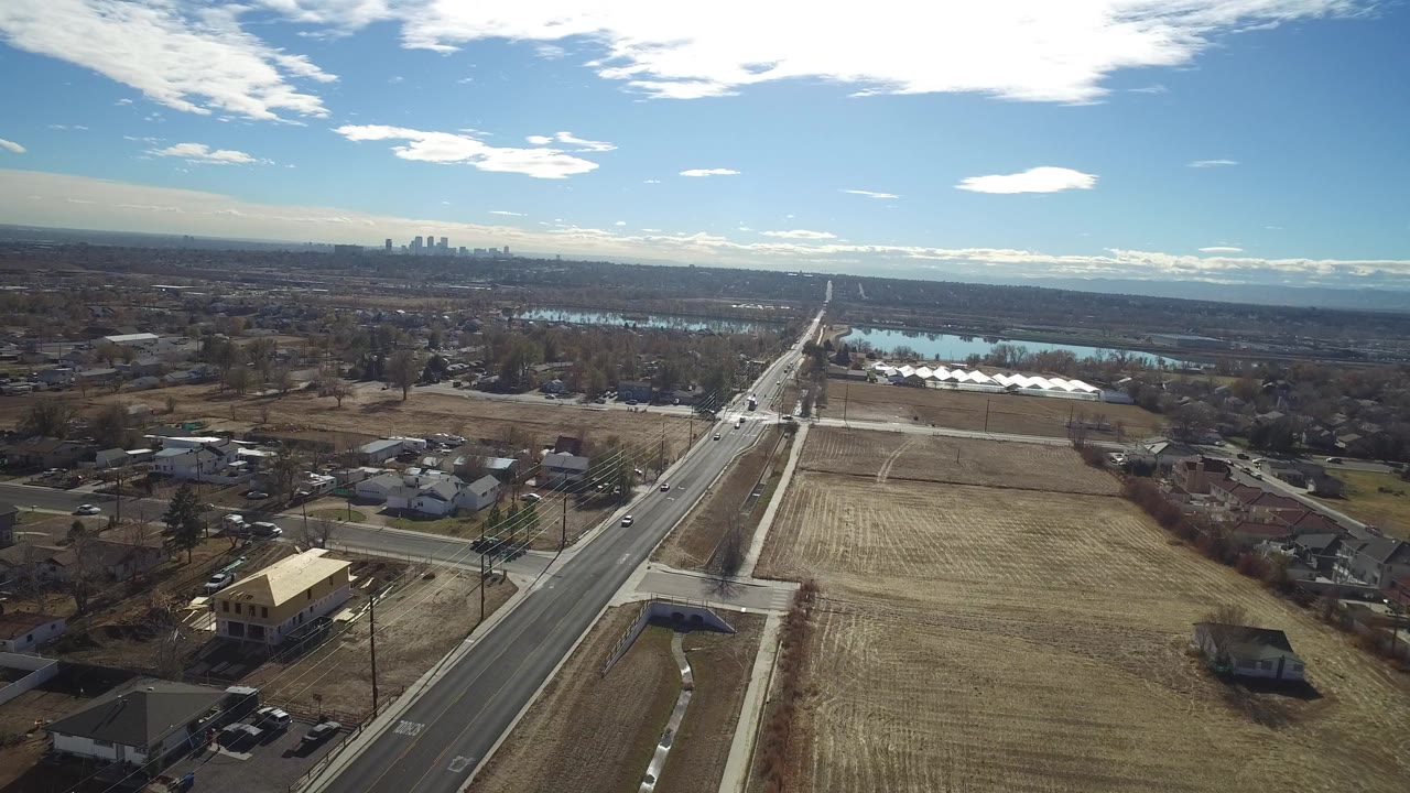 High aerial view in Westminster, Colorado, looking along a straight road past tan fields, suburban homes, a blue water body, distant skyline, and mountains, with no visible people.