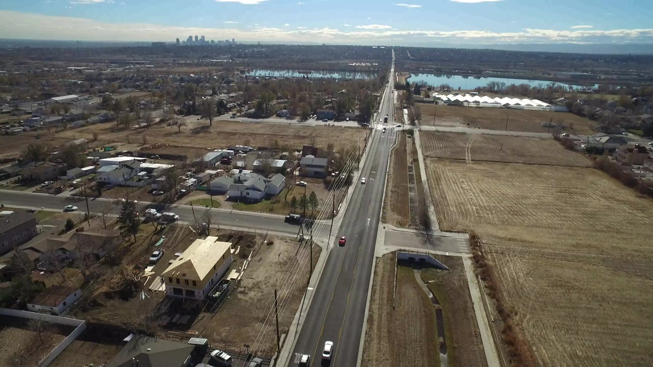High aerial view over Westminster, Colorado showing a straight road beside homes and dry fields, with water and a distant skyline on the horizon and no visible people.