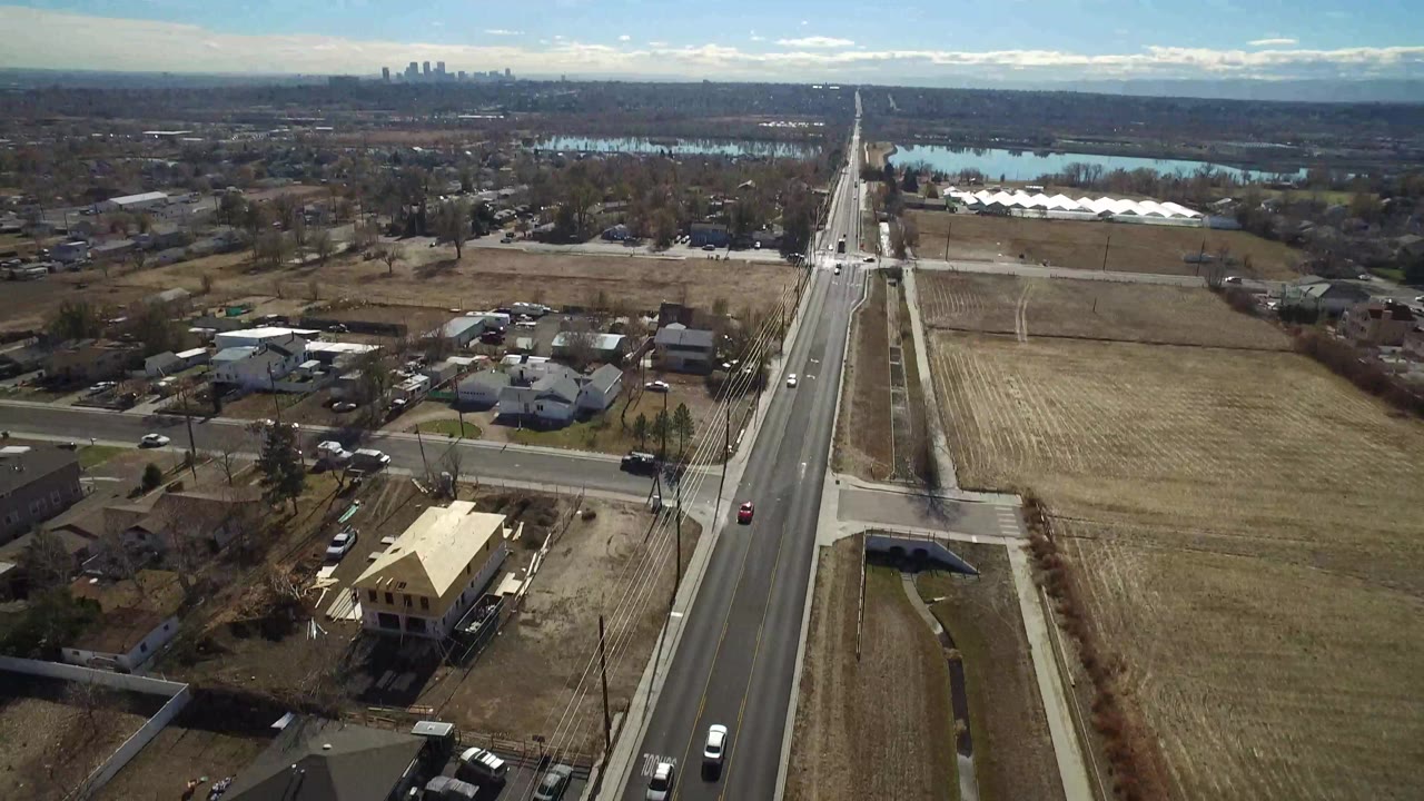 Aerial frame in Westminster, Colorado with houses and parked vehicles on the left, a straight road through the center, dry open field on the right, and no visible people.