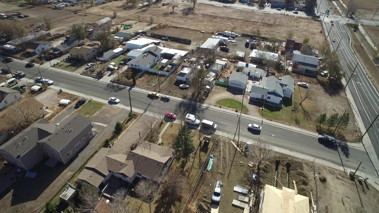 Aerial view in Westminster, Colorado showing the same residential street from slightly farther along, with houses, driveways, parked vehicles, dry lots, and no clearly visible people.