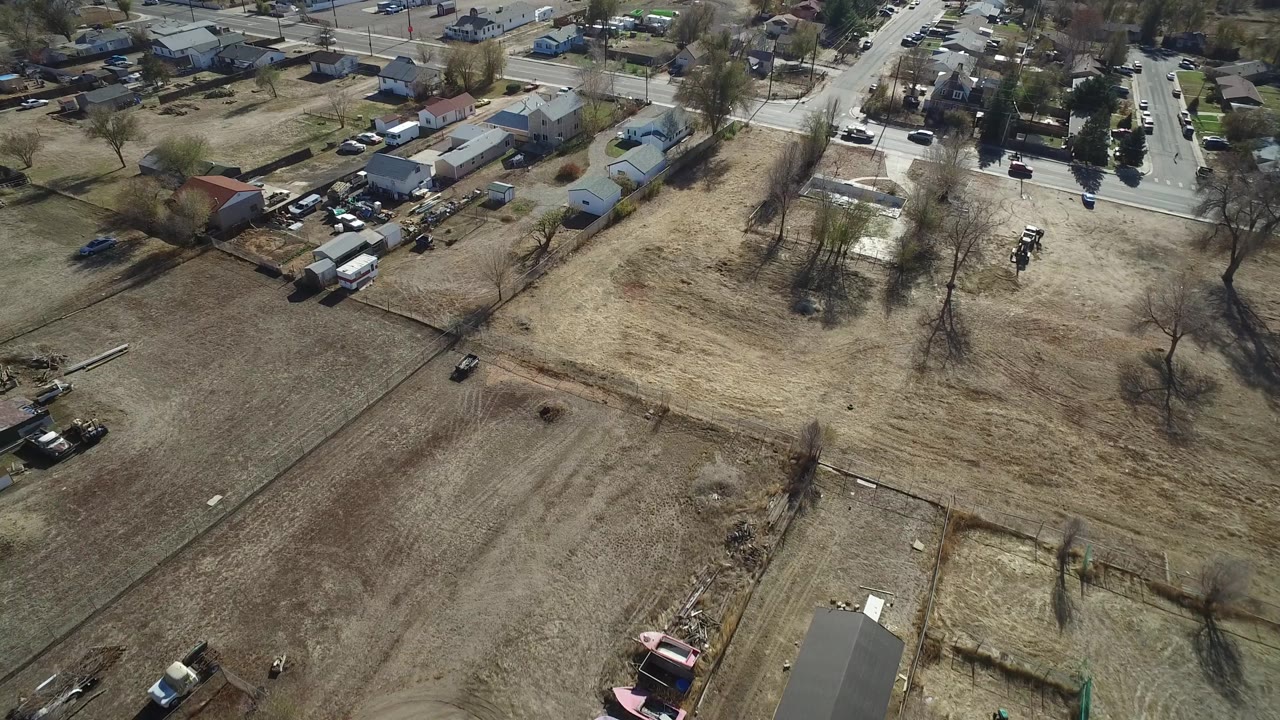 A slightly shifted aerial view of Westminster, Colorado showing a broad dry field in front, houses and outbuildings beyond it, and a paved road at the upper edge, with no visible people.