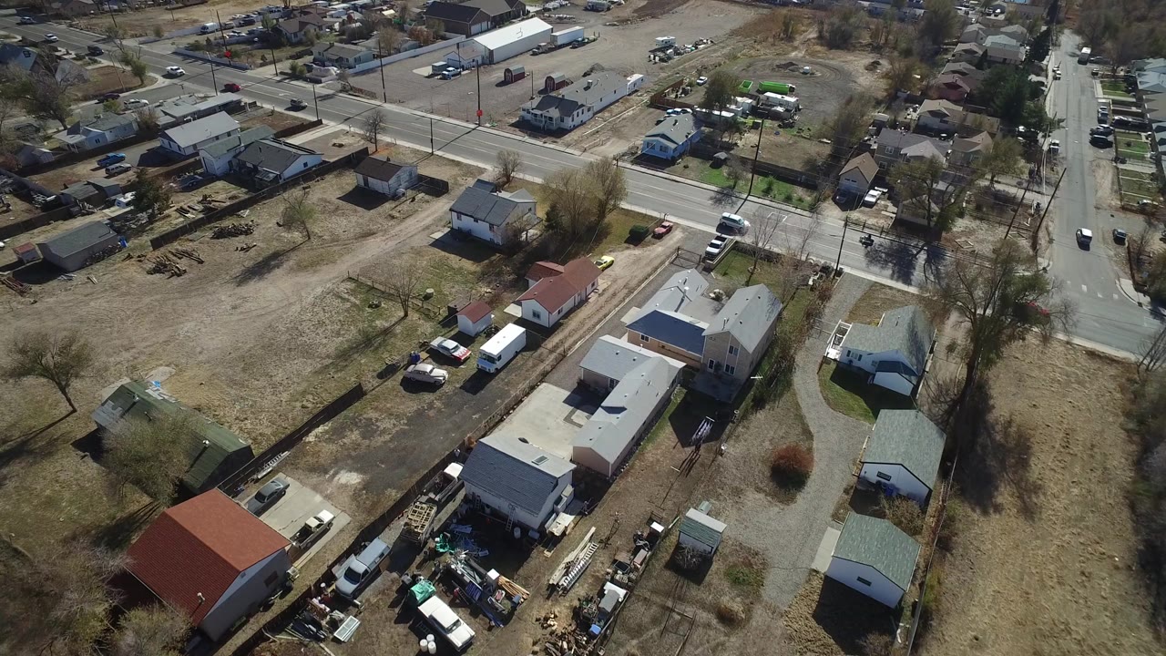 Drone view over Westminster, Colorado, with the road running near the top of frame, small houses and sheds below, open brown lots, parked vehicles, and no visible people.