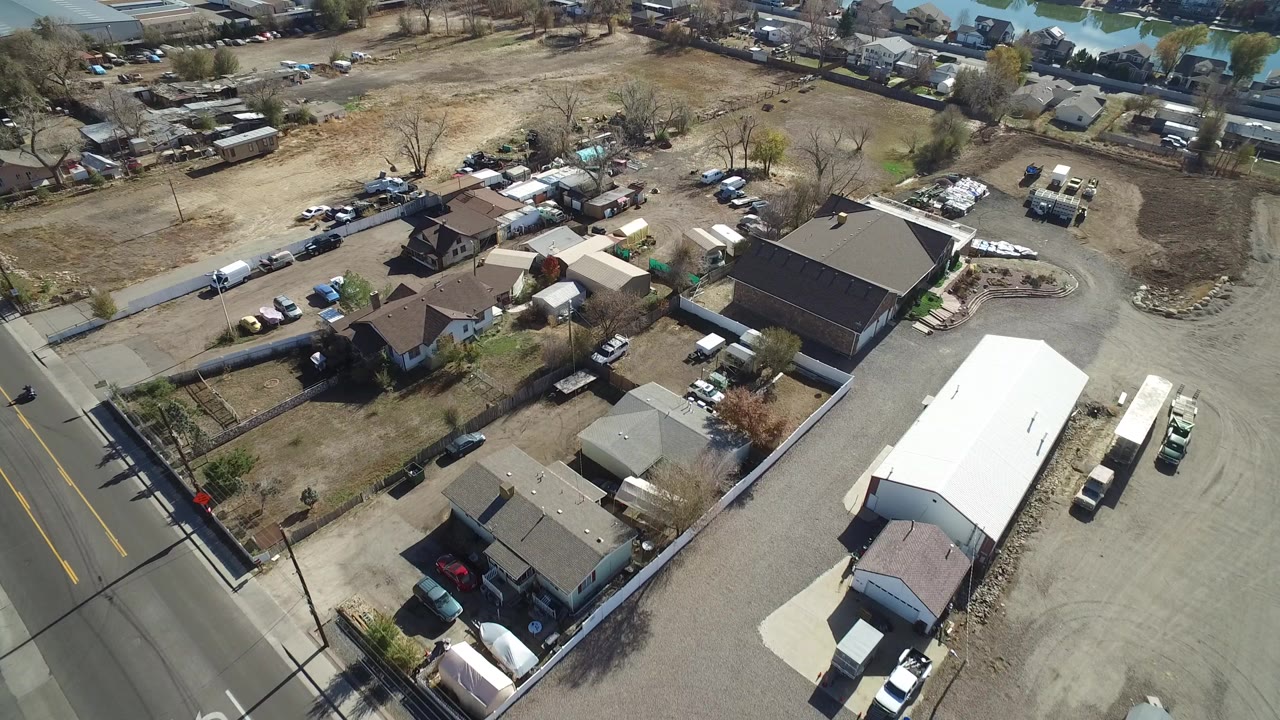 Aerial frame over Westminster, Colorado showing a road at left, detached homes, outbuildings, parked vehicles, dry open lots, and no visible people.