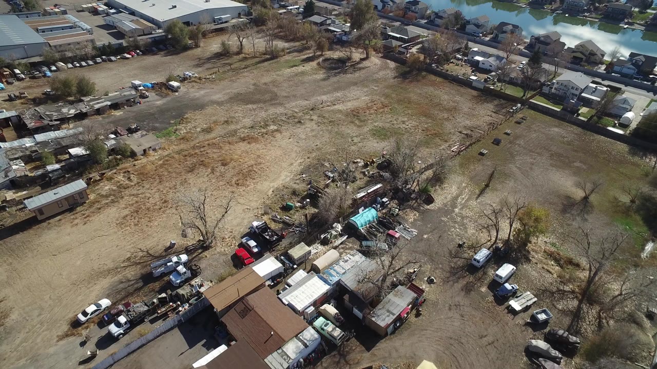 High aerial view over Westminster, Colorado showing a dry open lot, small buildings, parked vehicles, residential roofs, leafless trees, and no visible people.