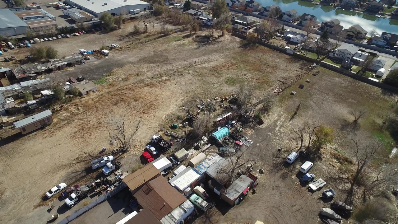 Drone-height view in Westminster, Colorado with the open dirt and grass parcel centered, clustered sheds and vehicles at lower left, houses and larger buildings beyond, and no visible people.