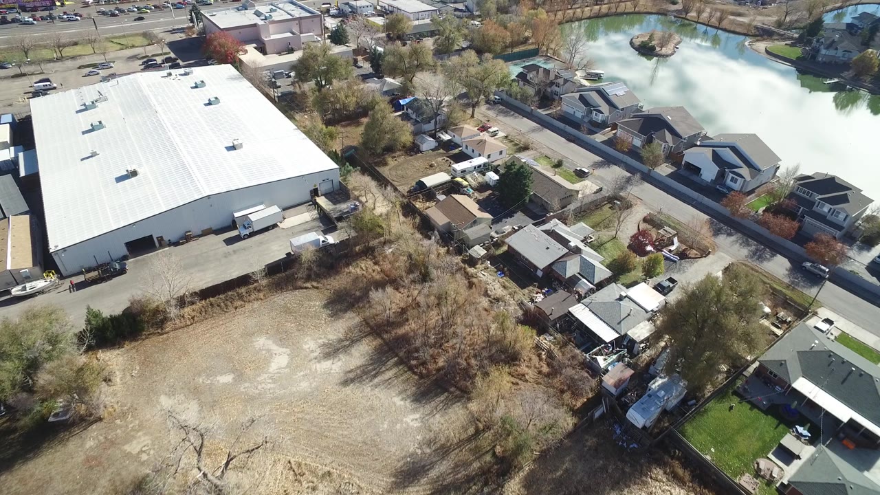Aerial frame in Westminster, Colorado with the warehouse roof on the left, fenced yards and houses below, a road through the middle, and pale water at the upper right, with no clearly visible people.