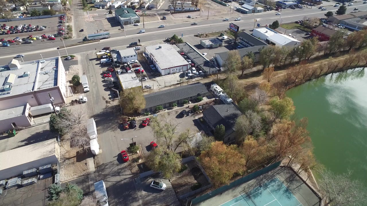 Aerial frame over Westminster, Colorado showing low-rise commercial buildings, parking lots, a road with vehicles, trees, and a green water feature, with no visible people.