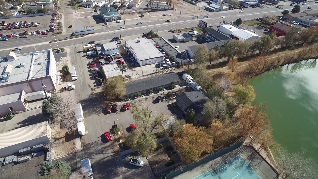 Aerial frame over Westminster, Colorado with the commercial roofs shifted across the foreground, parked cars along the lots, moving road traffic, trees, and green water, with no visible people.