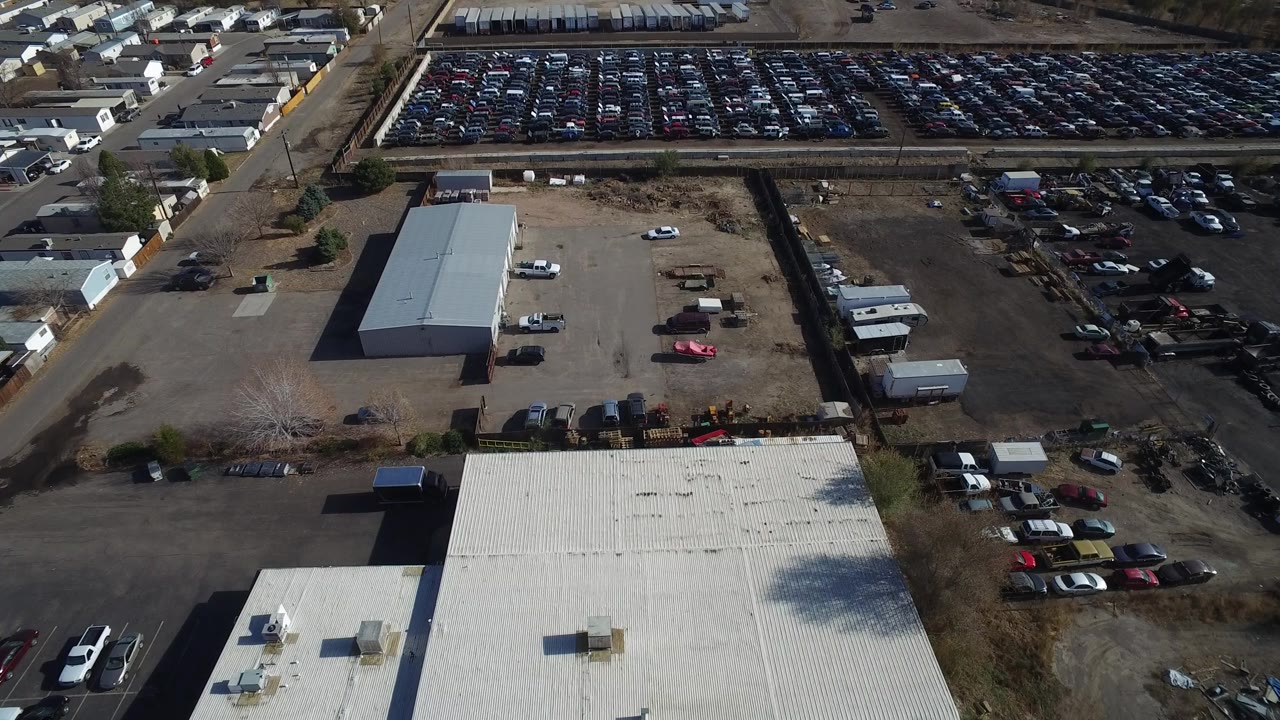 Opening aerial frame over an industrial area in Westminster, Colorado, with a white roof in the foreground, rows of parked cars beyond a road, and no visible people.