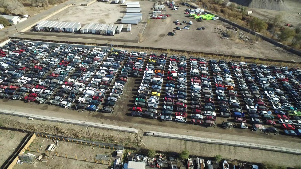 A high aerial frame over Westminster, Colorado shows dense rows of parked cars beside a straight road, with low industrial buildings and dry lots in the background and no visible people.