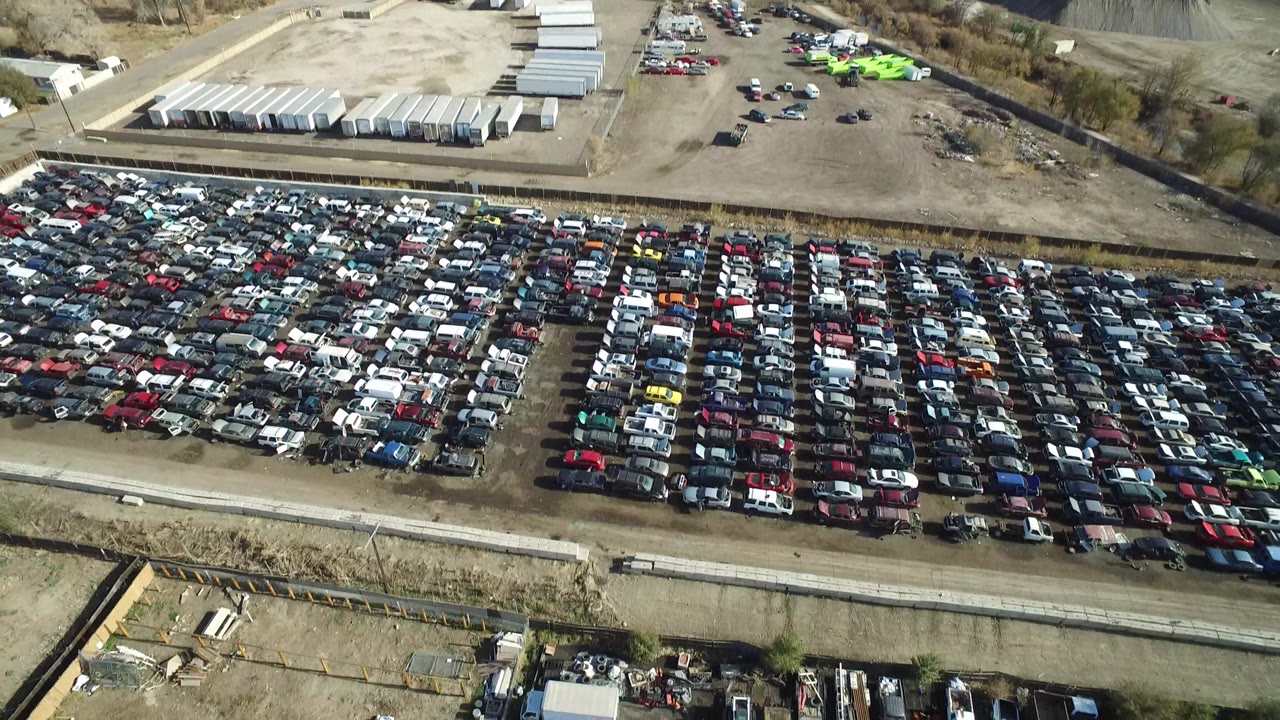 The second frame holds on the Westminster aerial view, with the car rows centered more tightly and the upper background showing storage buildings, parked trailers, and dry open ground with no visible people.