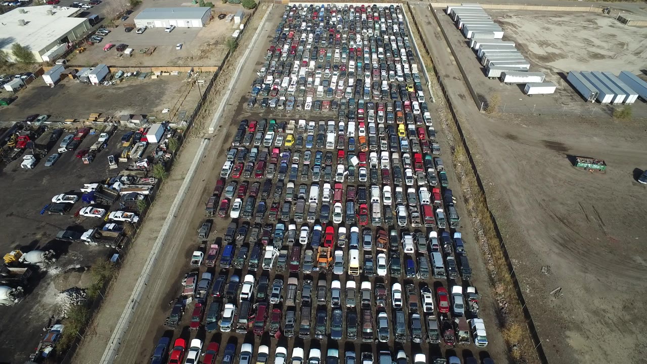 Opening aerial frame over Westminster, Colorado showing dense rows of parked cars beside a dirt road, gravel lots, low buildings, and no visible people.