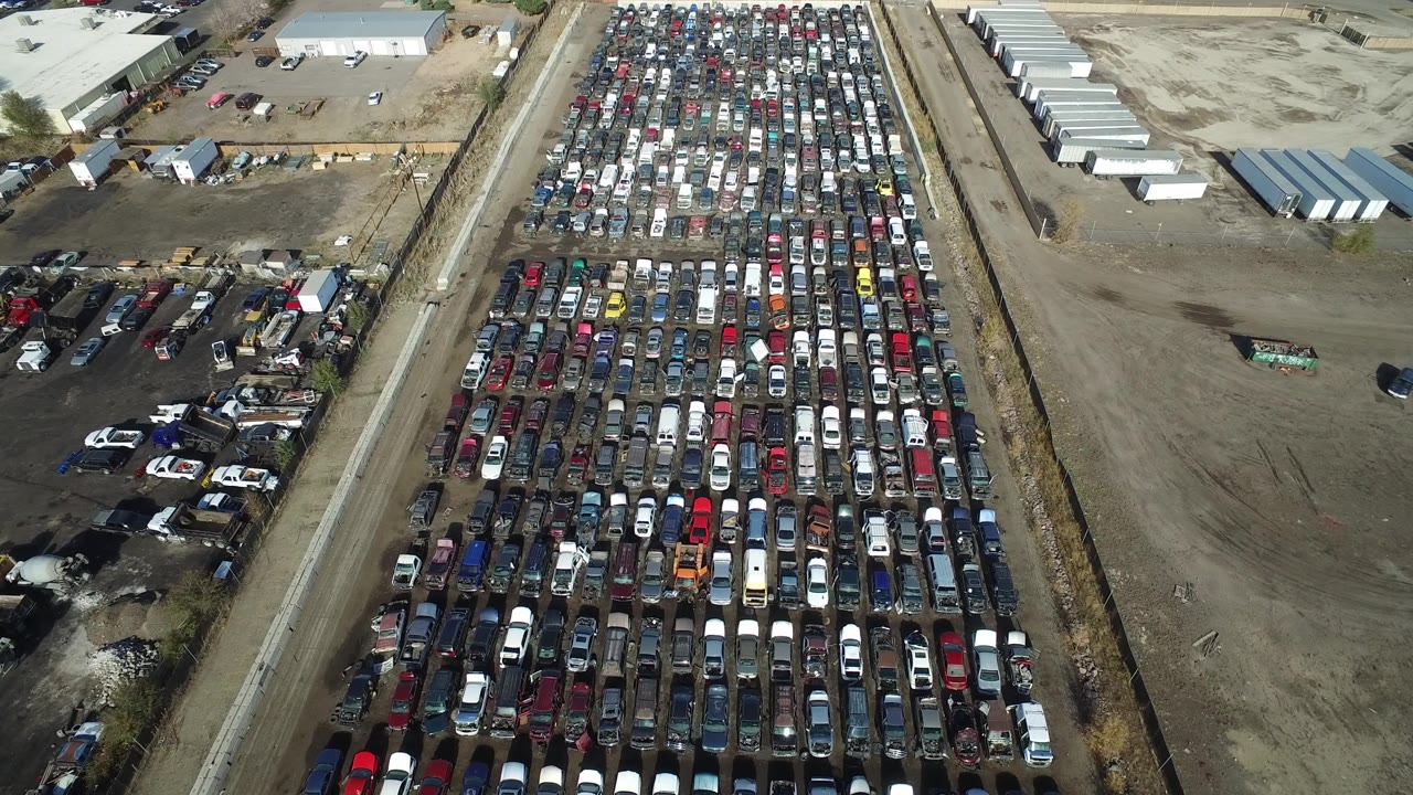 Aerial angle across parked cars, a dirt road, and sparse industrial lots.