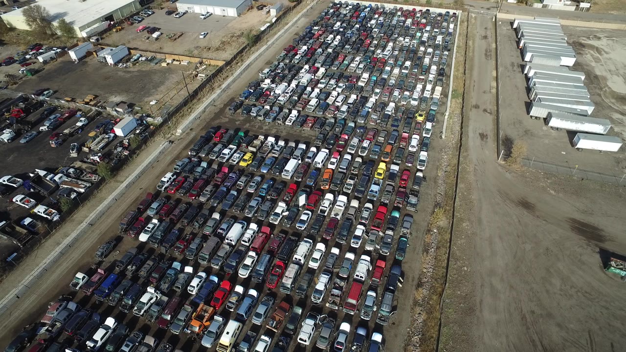 Aerial frame over a crowded vehicle lot in Westminster, Colorado, with dense rows of cars, dirt access roads, low industrial buildings, and no visible people.
